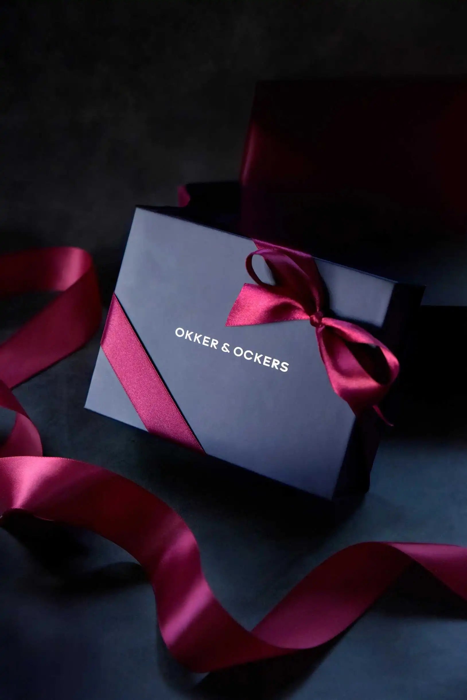 Okker & Ockers deep blue gift box with aubergine ribbon and silver logo, set against a dark background.