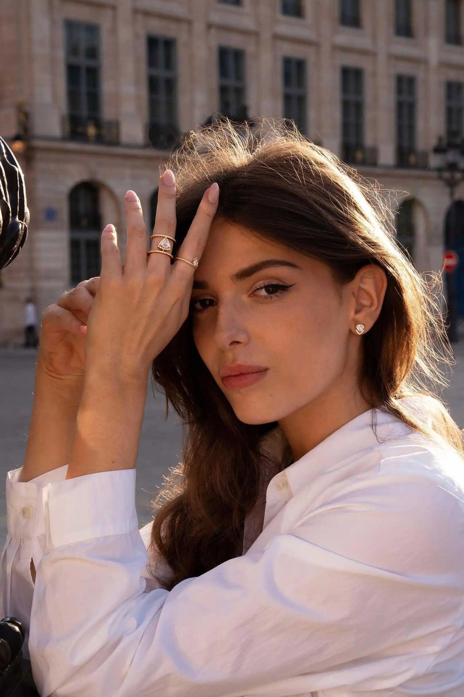 A model wearing the Audrey Icon ring, Victoria Accent ring, and Victoria Studs. The triangle-cut lab-grown diamonds catch the golden sunlight as she poses effortlessly in a crisp white shirt, with a Parisian backdrop in soft focus.