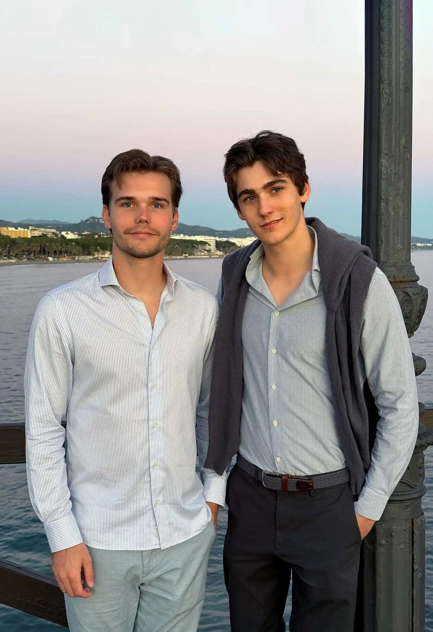 Brothers Ilan and Robin Okker standing together by the waterfront at sunset, dressed in light-striped shirts.