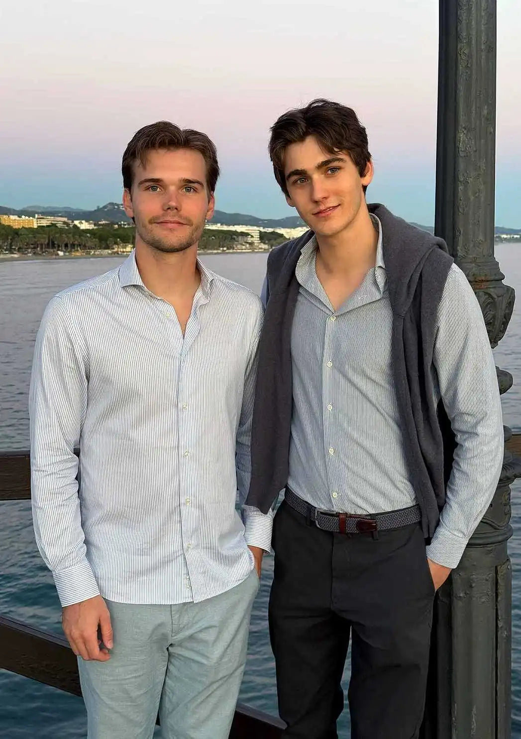 Brothers Ilan and Robin Okker standing together by the waterfront at sunset, dressed in light-striped shirts.