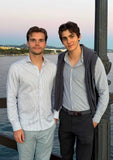 Brothers Ilan and Robin Okker standing together by the waterfront at sunset, dressed in light-striped shirts.